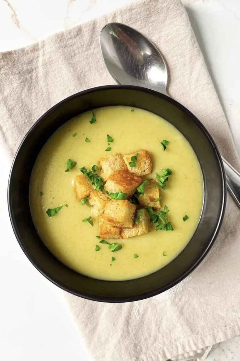 Un bol de sopa cremosa de crema de ajo porro con picatostes y hierbas, colocado en una servilleta con una cuchara al lado.