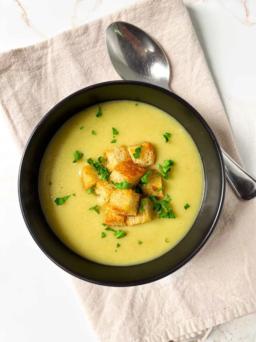 A bowl of creamy leek and potato soup topped with croutons and chopped herbs, next to a spoon on a beige napkin.