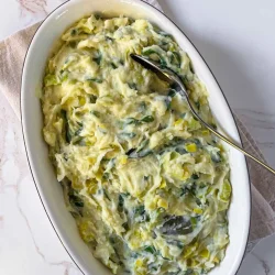 Oval dish of creamy mashed potatoes with leek and Swiss chard, served with a spoon on a light surface.