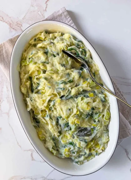 Oval dish of creamy mashed potatoes with leek and Swiss chard, served with a spoon on a light surface.