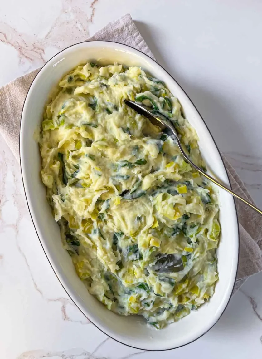 Plato ovalado de cremoso puré de papas mezclado con verduras y puerros picados, con una cuchara apoyada en el interior.