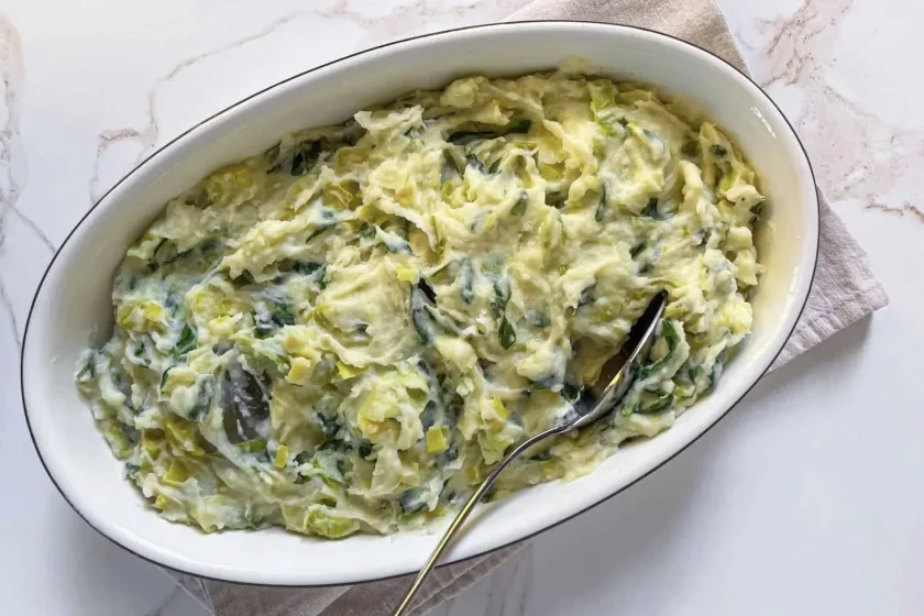 Plato ovalado de cremoso puré de papas con puerros y hierbas verdes, con una cuchara apoyada en su interior sobre un paño beige.