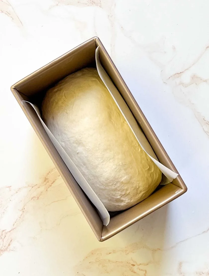 Una bola de masa en un molde para pan forrado con pergamino sobre una superficie de mármol claro.