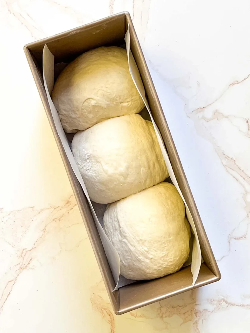 Tres bolas de masa en un molde rectangular forrado con papel pergamino, listas para hornear.