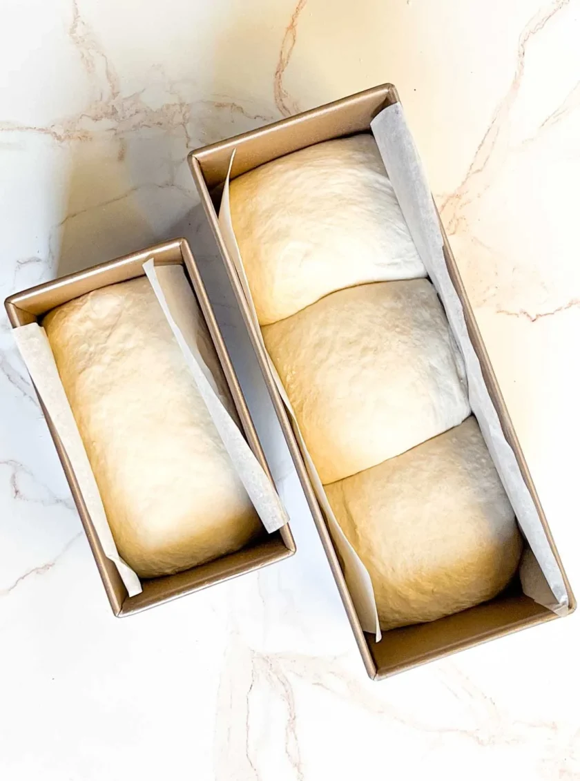 Dos moldes para pan con masa fermentada, uno grande con tres secciones y otro pequeño con una sola sección, sobre una superficie de mármol.