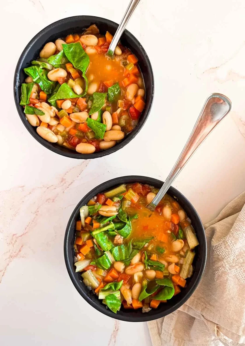 Two bowls of vegetable and bean soup with leafy greens, on a light surface with a beige napkin.