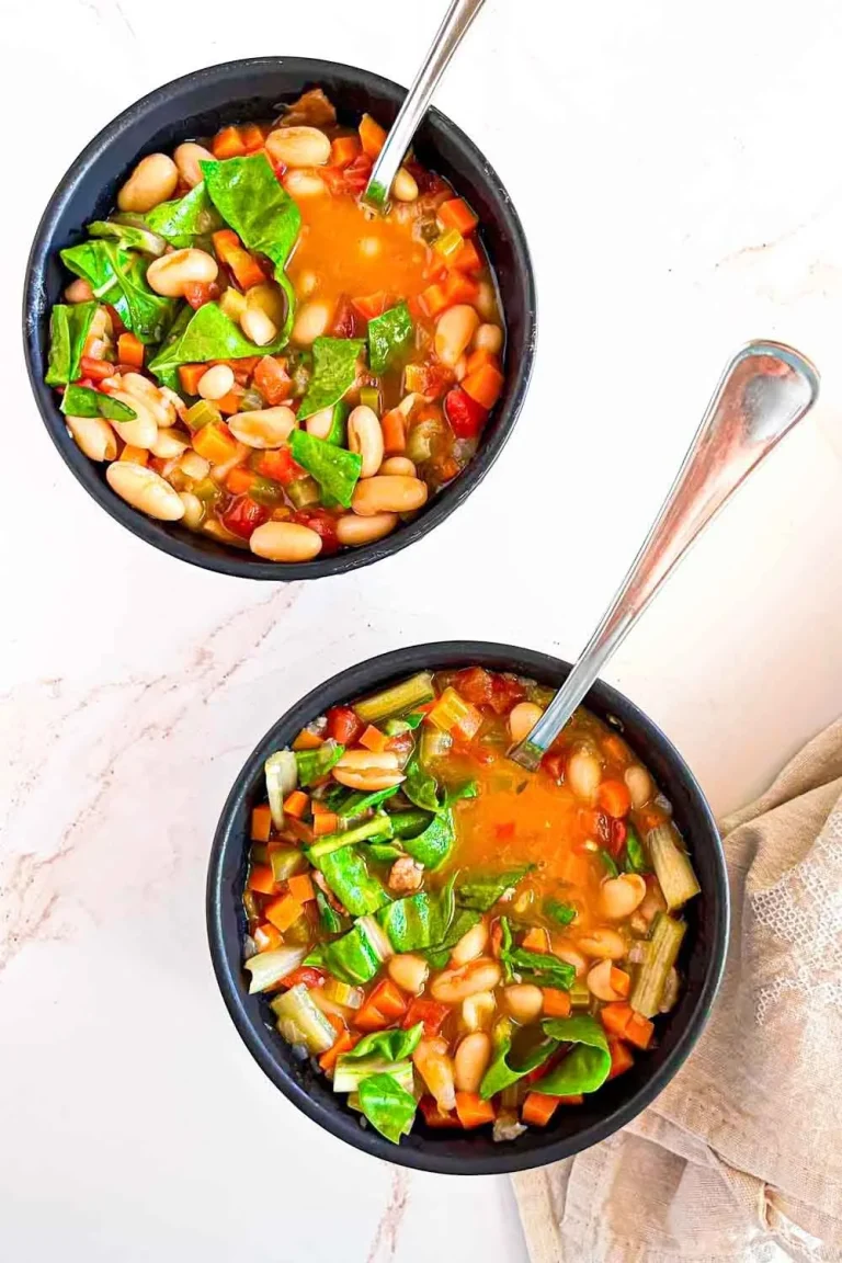 Dos cuencos de Menestra de alubias con acelgas, rellenos de espinacas, zanahorias y tomates, se asientan sobre una superficie blanca, una deliciosa versión de la receta de sopa de alubias con acelgas.