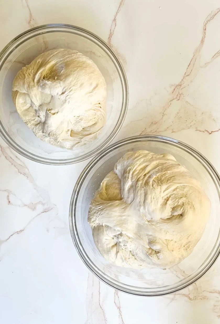 Dos cuencos de cristal llenos de masa fermentada sobre una superficie de mármol blanco.