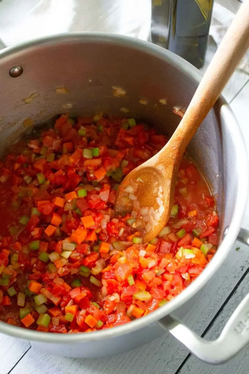 Verduras cortadas en dados que se cocinan en una olla con una cuchara de madera, incluyendo tomates, zanahorias, apio y cebollas. Todo para hacer sopa de lentejas a la italiana.
