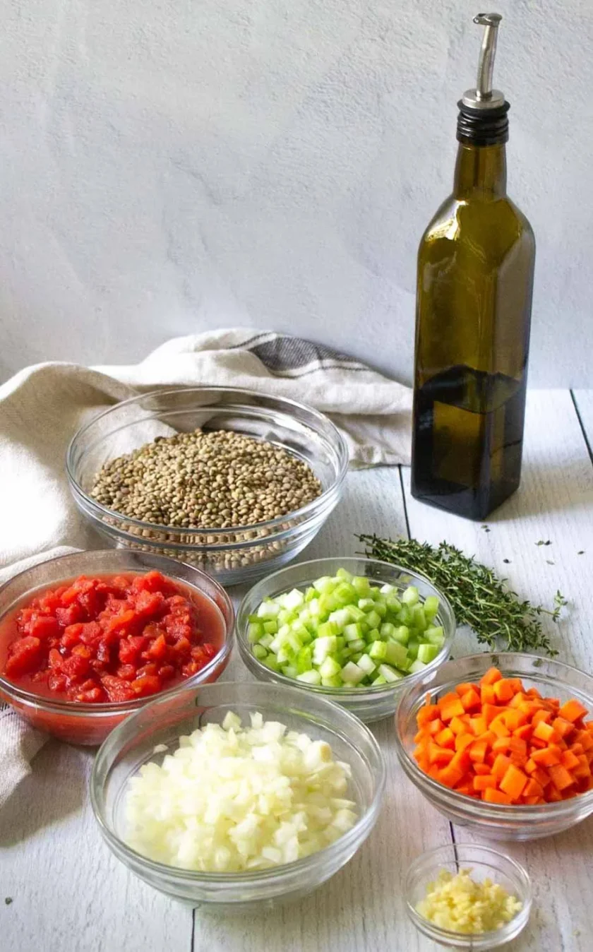 Cebollas picadas, zanahorias, apio, tomates, lentejas, ajo, tomillo fresco y aceite de oliva sobre una mesa blanca. Estos son los ingredientes para hacer sopa de lentejas a la italiana.