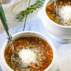 Dos cuencos blancos de sopa de lentejas cubiertos de queso parmesano rallado, cada uno con una cuchara, sobre una superficie de madera clara.
