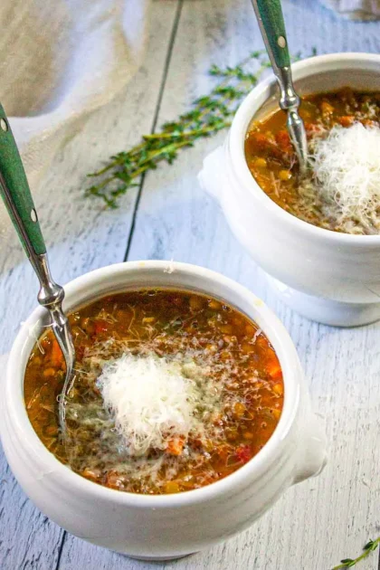 Dos cuencos blancos de sopa de lentejas cubiertos de queso parmesano rallado, cada uno con una cuchara, sobre una superficie de madera clara.