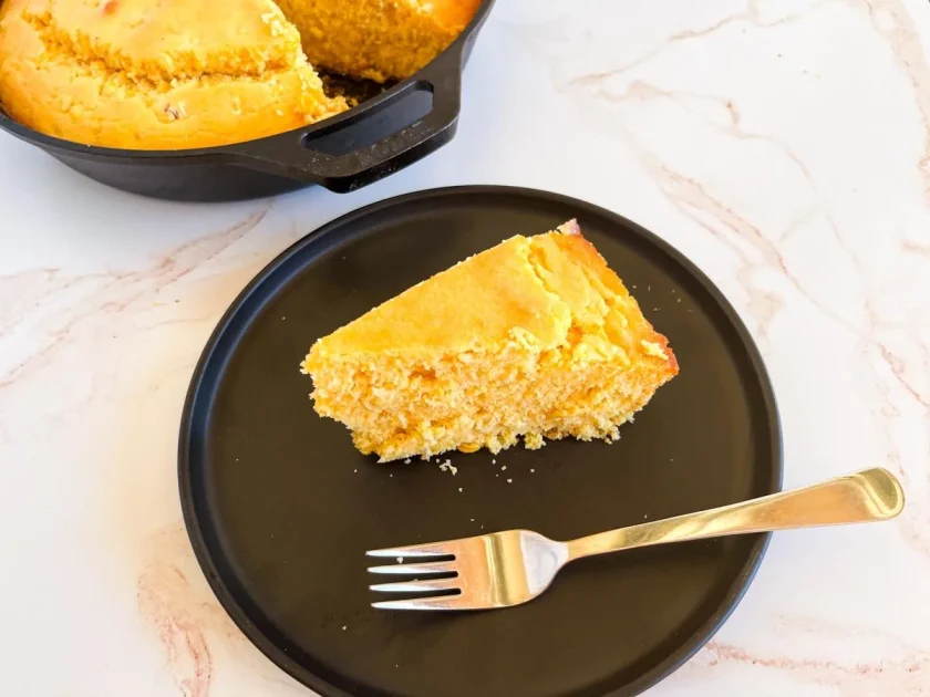Una rebanada de pan de maíz en un plato negro con un tenedor, junto a una sartén de pan de maíz en una superficie blanca.