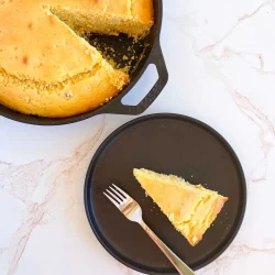 Una sartén de pan de maíz con una rebanada cortada y servida en un plato negro con un tenedor.