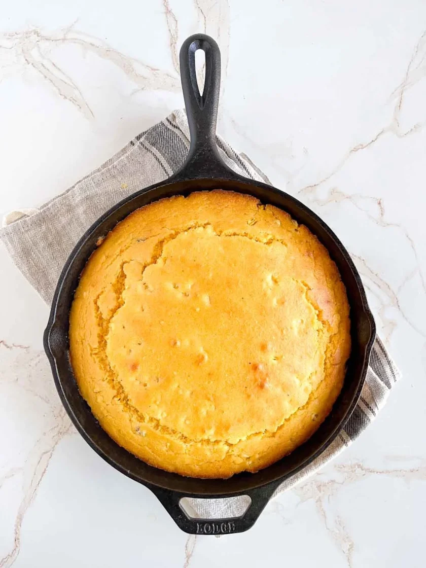 Pan de maíz dorado horneado en una sartén de hierro fundido negro, descansando sobre un paño de cocina gris en una superficie de mármol.