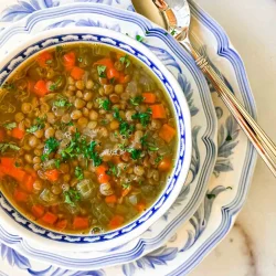 A bowl of lentil soup with carrots and herbs, served on a patterned plate with a spoon beside it.
