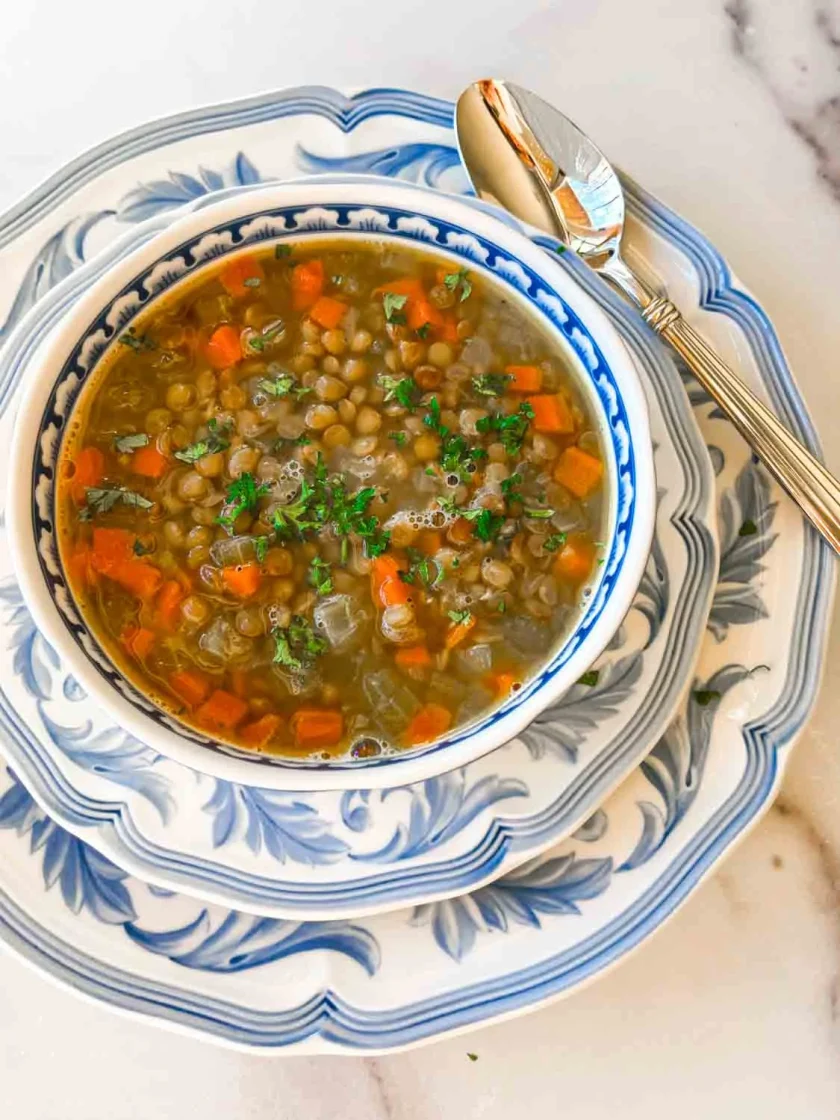 Un cuenco de sopa de lentejas con zanahorias y hierbas, servido en un plato azul y blanco con una cuchara al lado.