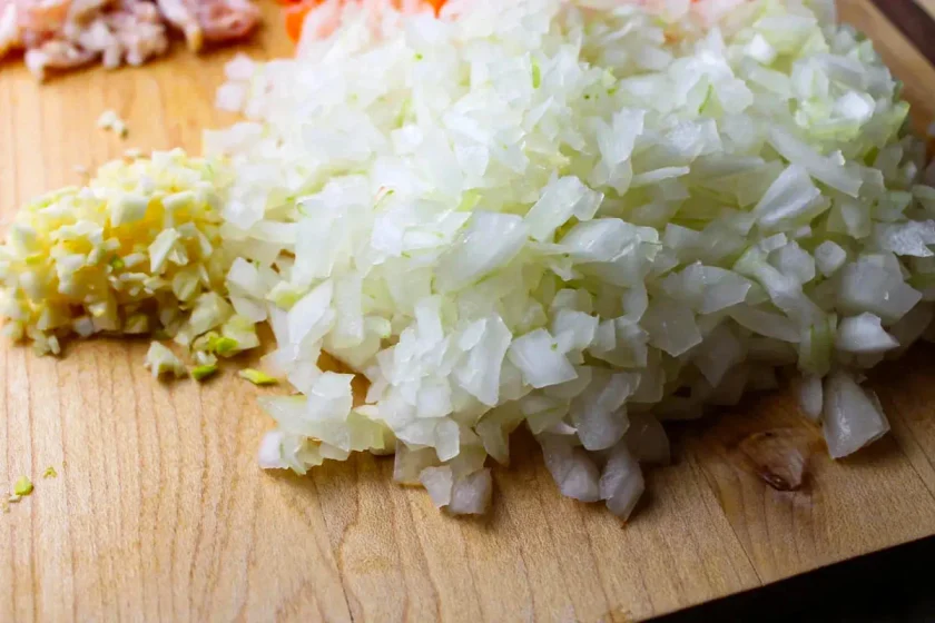 Cebollas picadas y ajo picado sobre una tabla de cortar de madera con otros ingredientes cortados en dados al fondo.