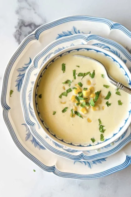 A bowl of creamy sopa, inspired by receta de crema de jojoto, is topped with corn and herbs on a blue and white patterned plate with a spoon.