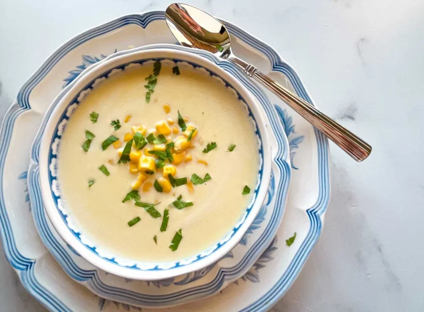 Tazón de sopa cremosa de maíz con guarnición de maíz y hierbas, en platos apilados con una cuchara al lado.