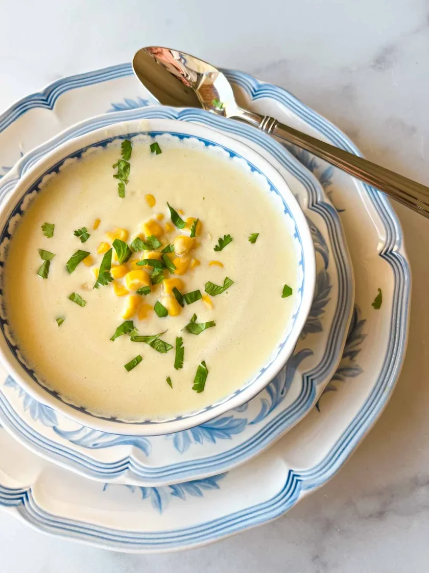 Tazón de sopa cremosa de maíz adornado con perejil, servido en platos azules y blancos con una cuchara.