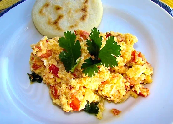 Perico de huevos revueltos con tomates y hierbas, adornados con cilantro, servidos con una arepa redonda pequeña en un plato blanco.