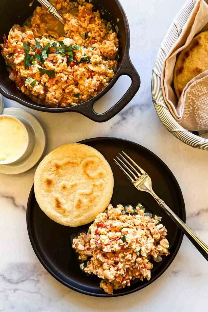 A plate with perido (scrambled eggs with sofrito,) an arepa, a fork, and a pan of perico with arepas on the side.