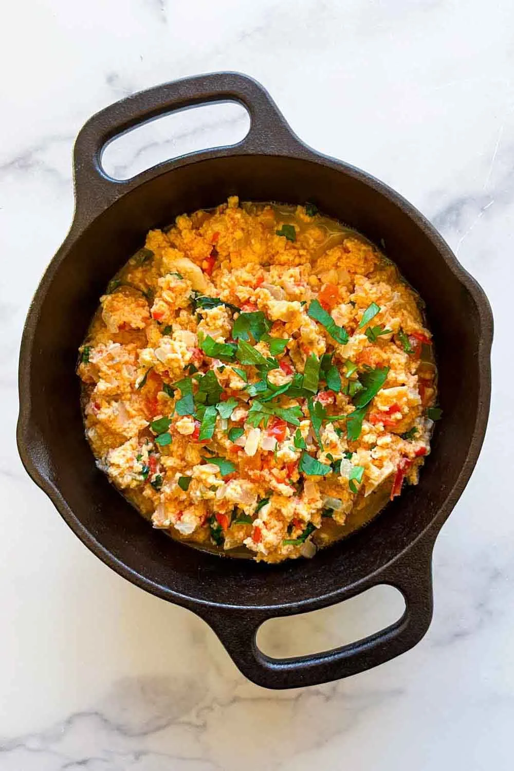 Huevos revueltos con tomates y hierbas en una sartén negra de hierro fundido sobre una superficie de mármol blanco.