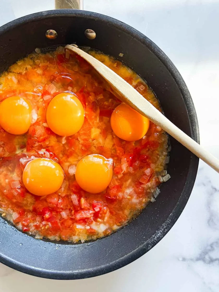 Five raw eggs on top of chopped tomatoes and onions cooking in a black pan with a wooden spoon.