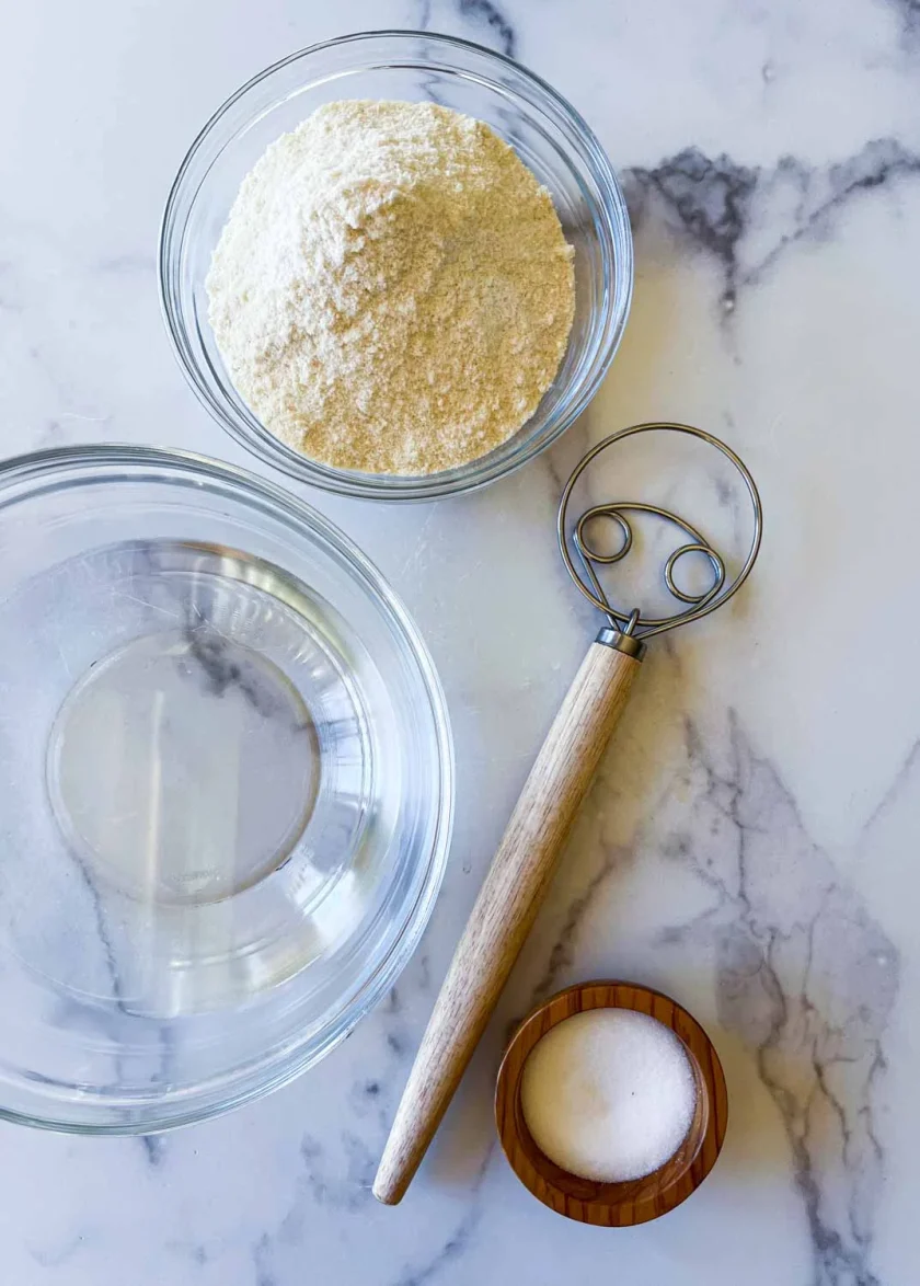 Ingrerdienets para hacer arepas> agua. harina de arepas y sal, además de un batidor da´s, en una superficie de mármol blanco.