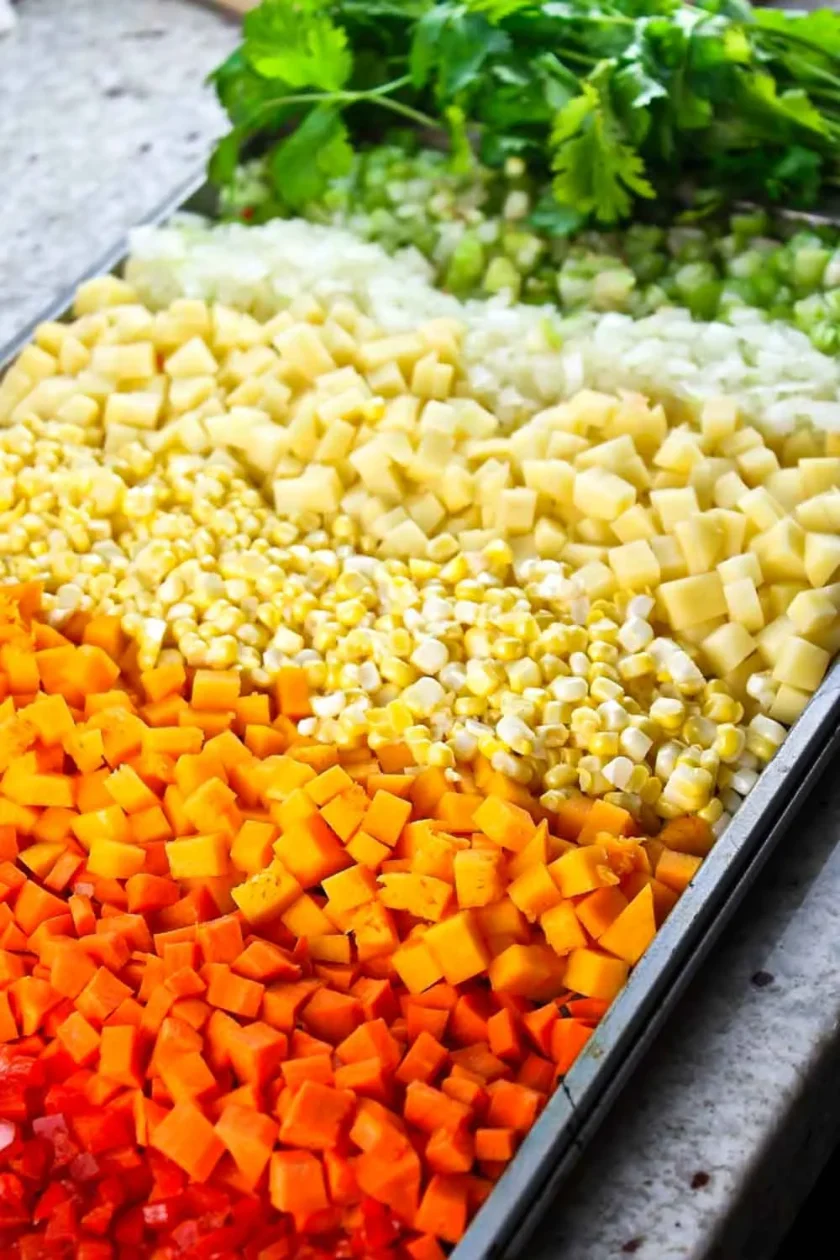Una bandeja con hileras de verduras troceadas: pimientos rojos, zanahorias, maíz, patatas, apio y cilantro.