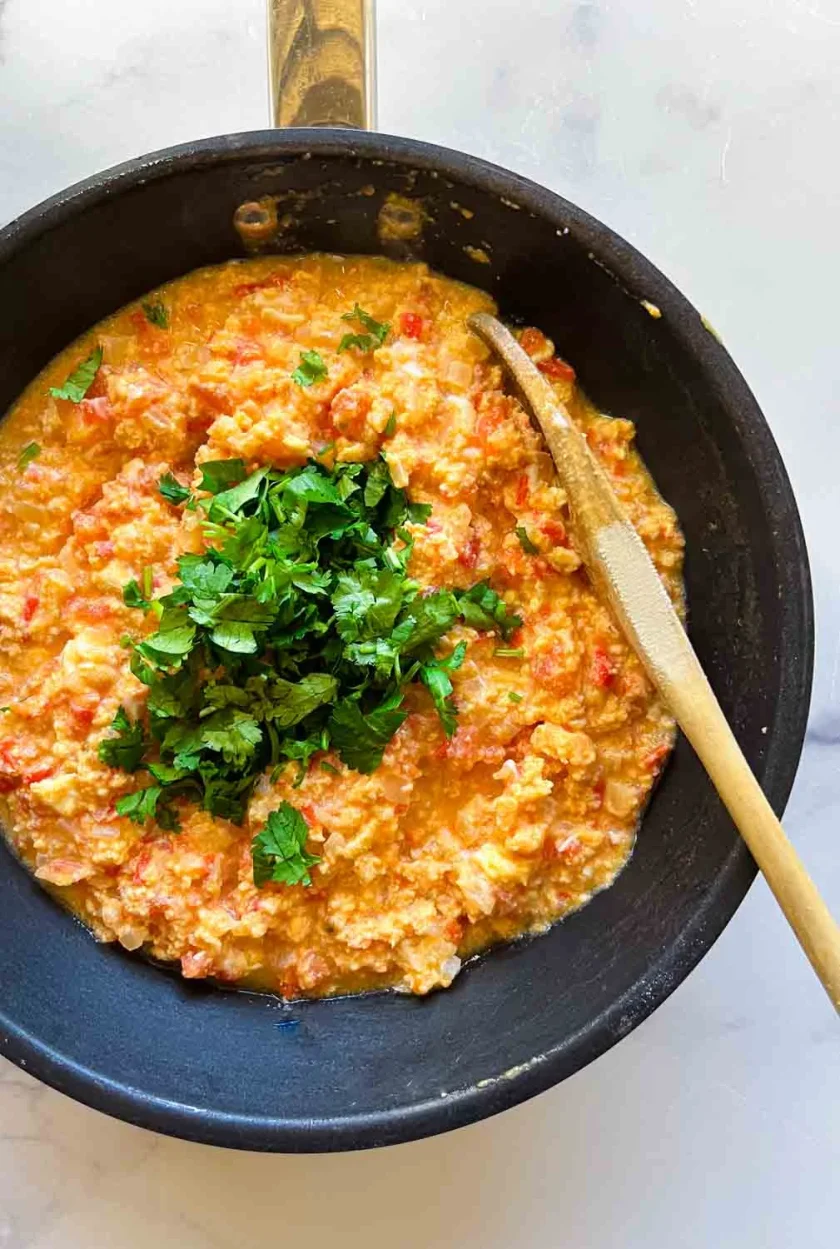A skillet of perico scrambled eggs with tomatoes, topped with fresh chopped herbs and a wooden spoon on the side.