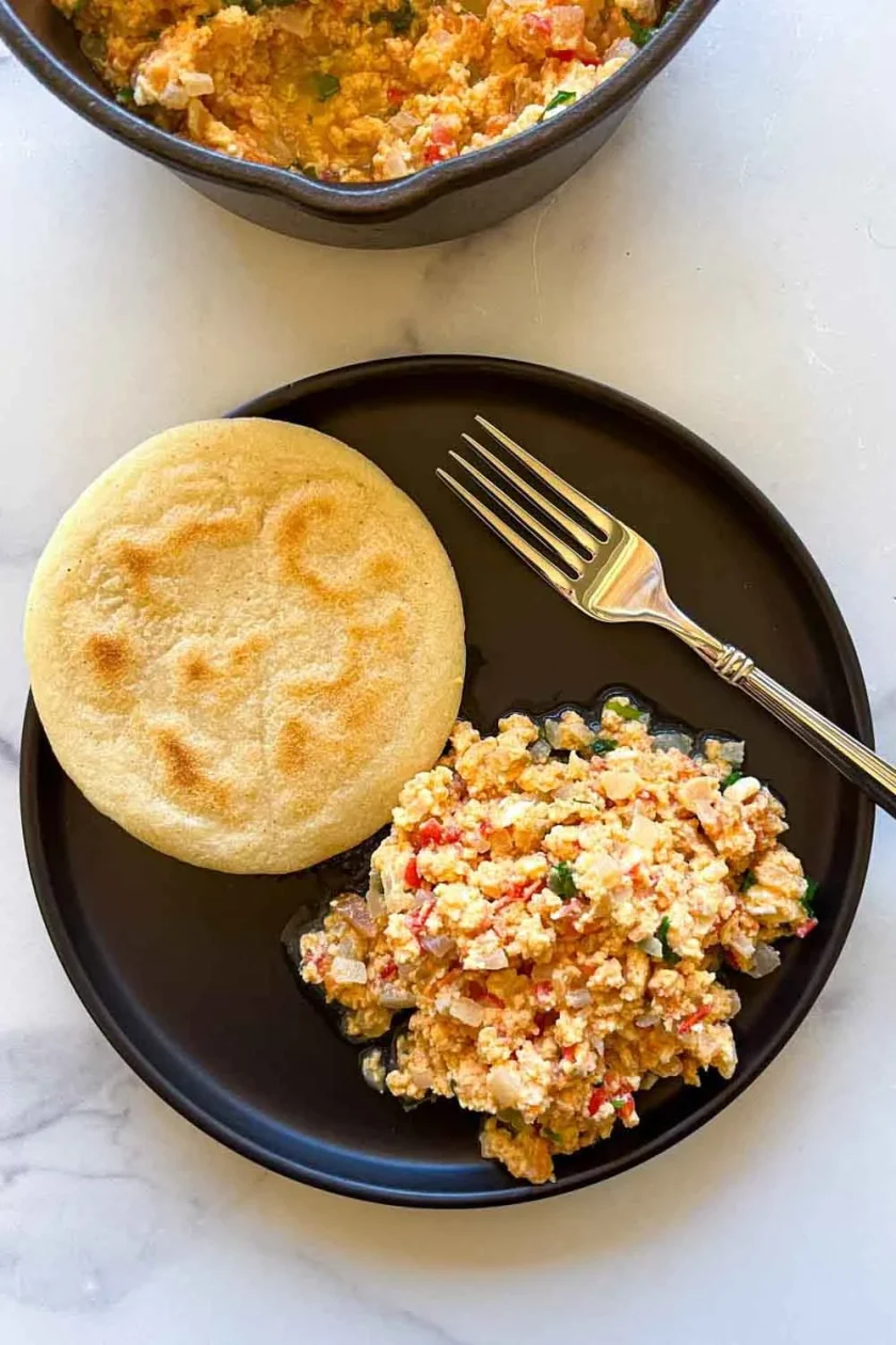Black plate with scrambled eggs and vegetables next to a round arepa, with a fork beside the food.