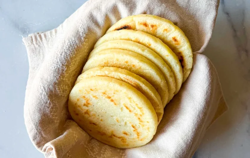 Seis arepas en un recipiente cubierto con una toalla de lino beige, sobre una superficie de mármol blanco.