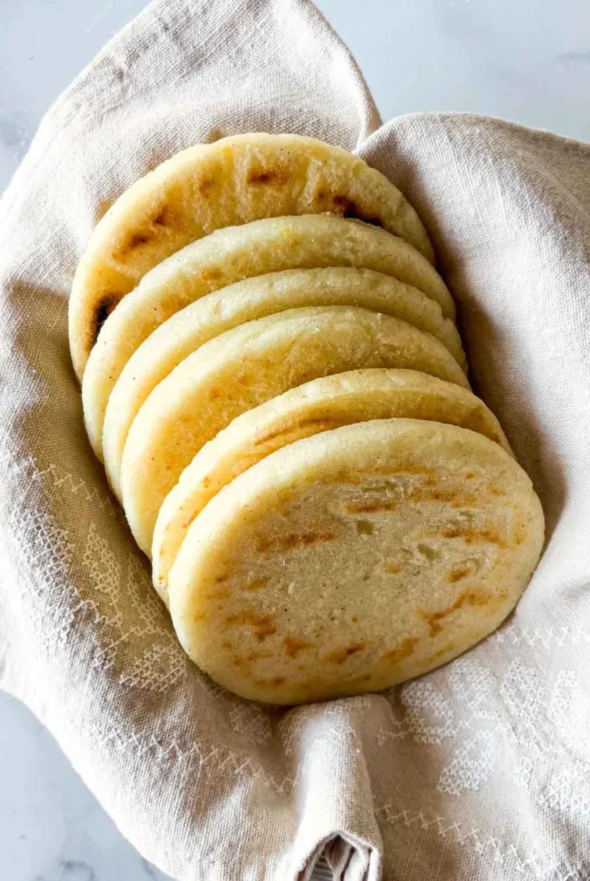 Seis arepas en un recipiente cubierto con una toalla de lino beige, sobre una superficie de mármol blanco.