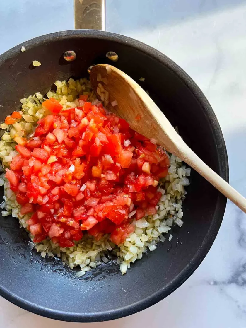 Diced onions and tomatoes in a skillet with a wooden spoon on a marble surface.