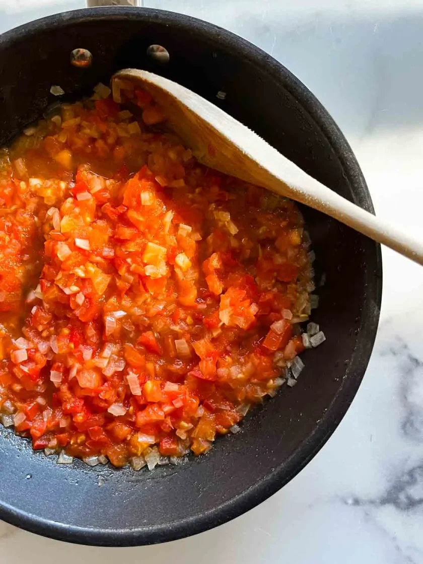 Chopped tomatoes and onions cooking in a black pan with a wooden spoon on a marble countertop.