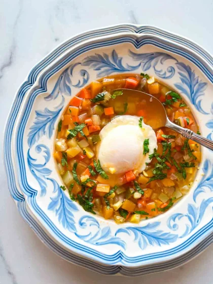 Un cuenco de sopa de verduras con un huevo escalfado encima, servido en un plato con motivos azules y blancos.