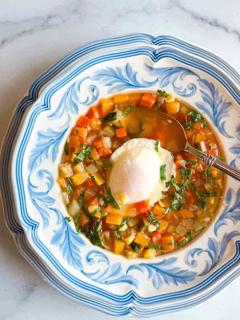 Un cuenco de sopa de verduras con un huevo escalfado y hierbas, servido en un plato decorativo azul y blanco.