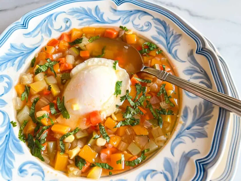 Colorida sopa de verduras con cilantro picadas y un huevo escalfado en un cuenco decorativo, con una cuchara al lado.