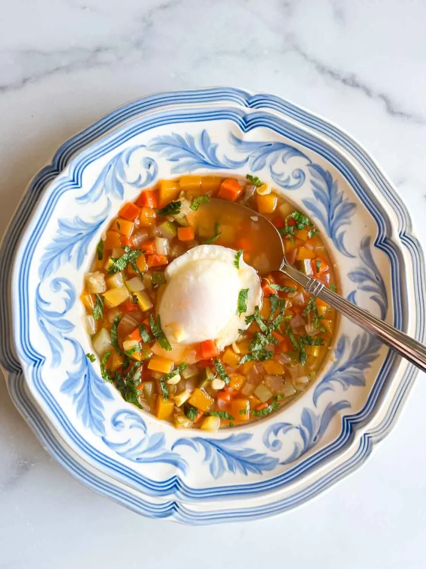 Un cuenco de sopa de verduras con un huevo escalfado, servido en un plato con motivos azules y blancos.