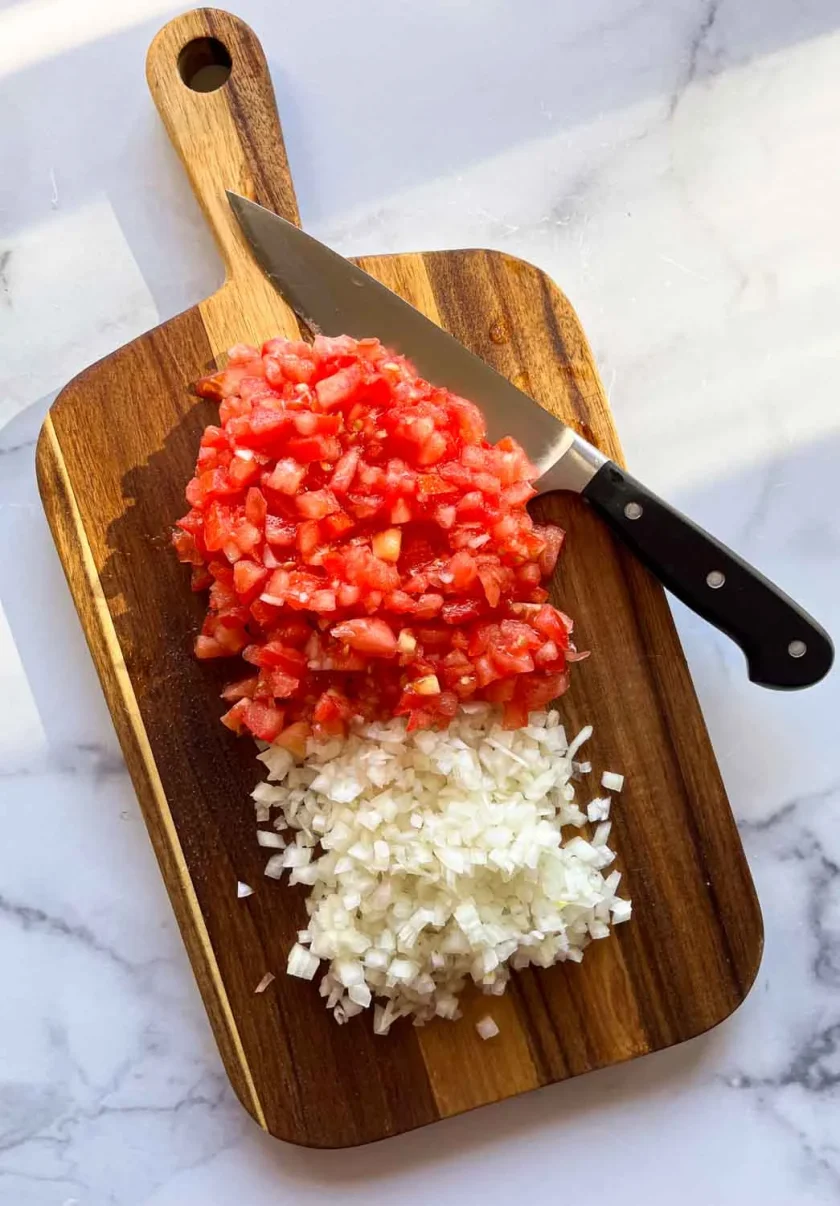 Chopped tomatoes and onions on a wooden cutting board with a knife beside them.