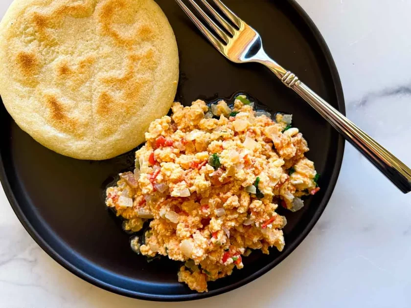 Perico: scrambled eggs with veggies and an arepa served on a black plate with a fork.
