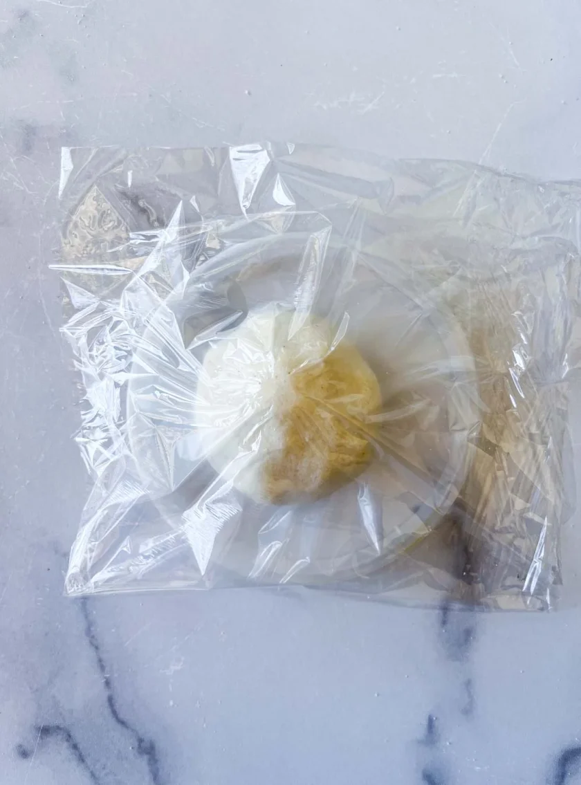 una bolita de masa de arepa sobre una tapa plástica. La tapa esta cubierto con film plástico y la bolita de masa también. Todo esta sobre una superficie de mármol blanco.