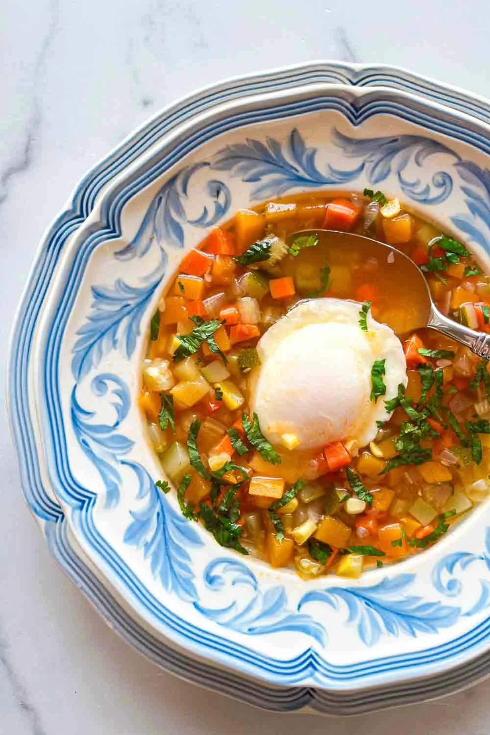 Un bol de sopa de verduras con huevo y cilantro - receta venezolana, con un huevo escalfado y hierbas frescas, servido en un plato decorativo azul y blanco.