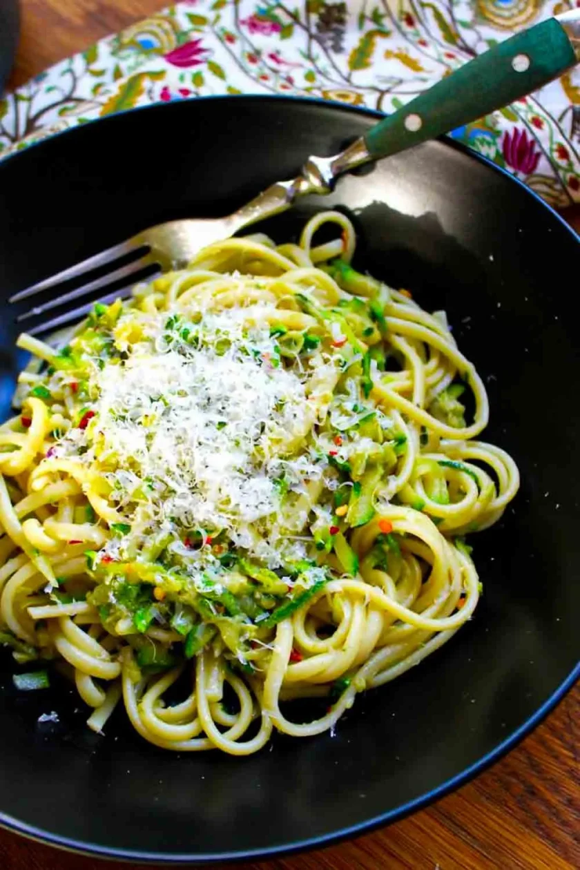 Un plato negro con linguini con calabacín y anchoa, servidos con queso parmesano. El plato tiene un tenedor.