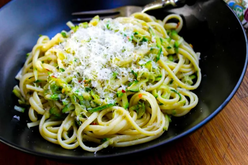 Un plato negro con linguini con calabac'in y anchoa, servidos con queso parmesano. El plato tiene un tenedir.