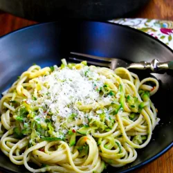 A black bowl of pasta with zucchini and anchovies asprinkled with Parmesan cheese, over a wooden surface.