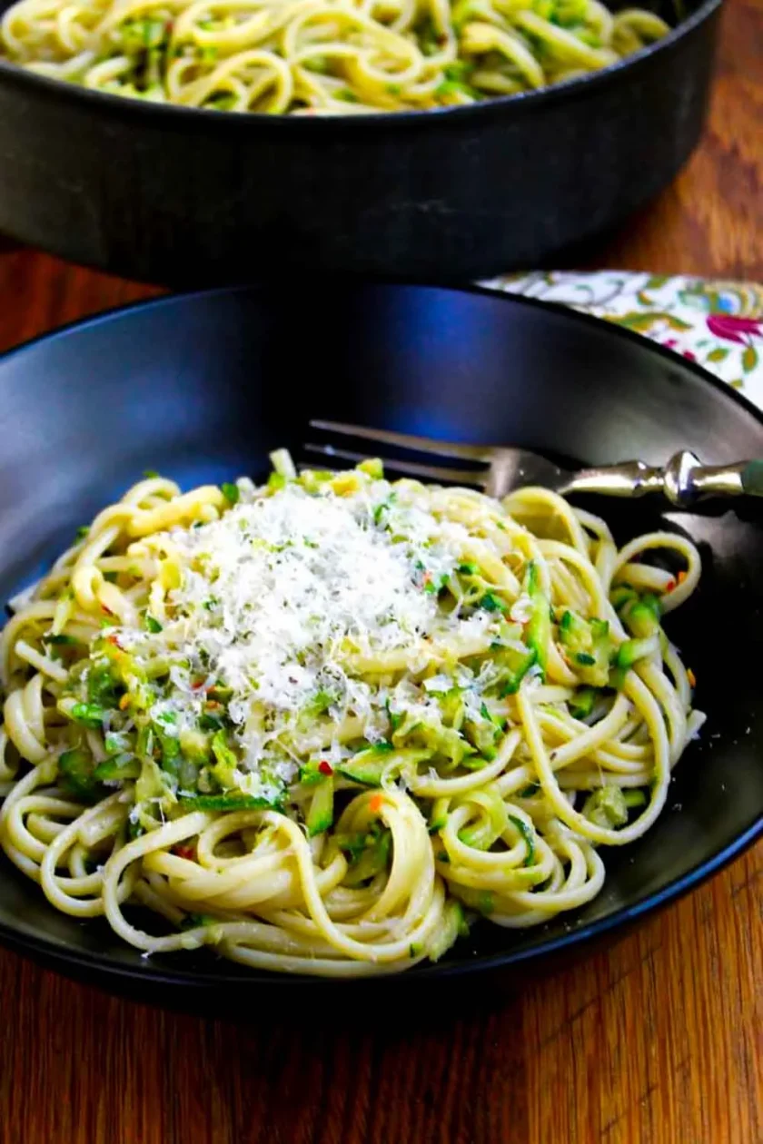 Un plato negro con linguini con calabac'in y anchoa, servidos con queso parmesano. El plato tiene un tenedir y al fono se ve el sartén con más pasta.