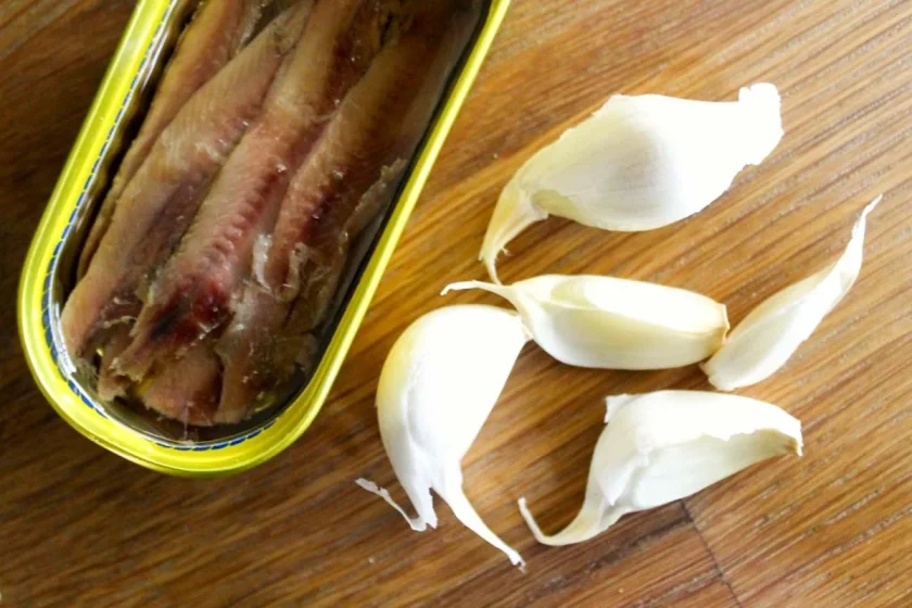 An open can of achovy filletes in olive oil and five garlic cloves on a wooden surface.